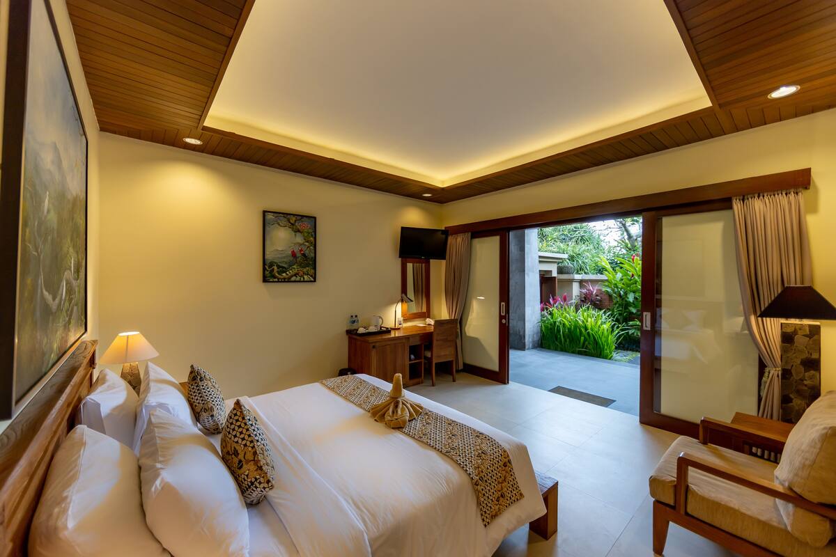 A hotel room with a large bed, a desk, a chair, and a view of a garden through a glass door.