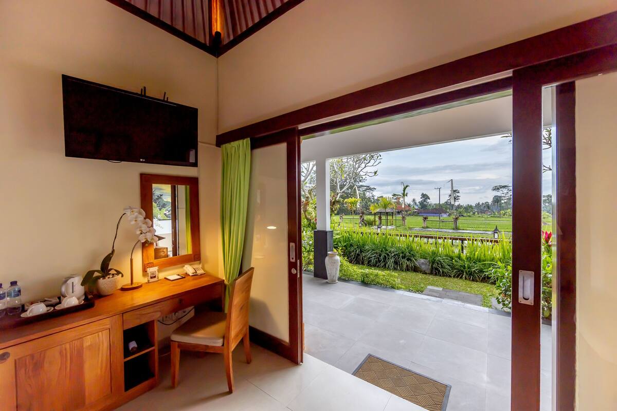 A room with a desk, chair, and a television mounted on the wall. There is a door leading to an outdoor area with a view of greenery and a pathway.