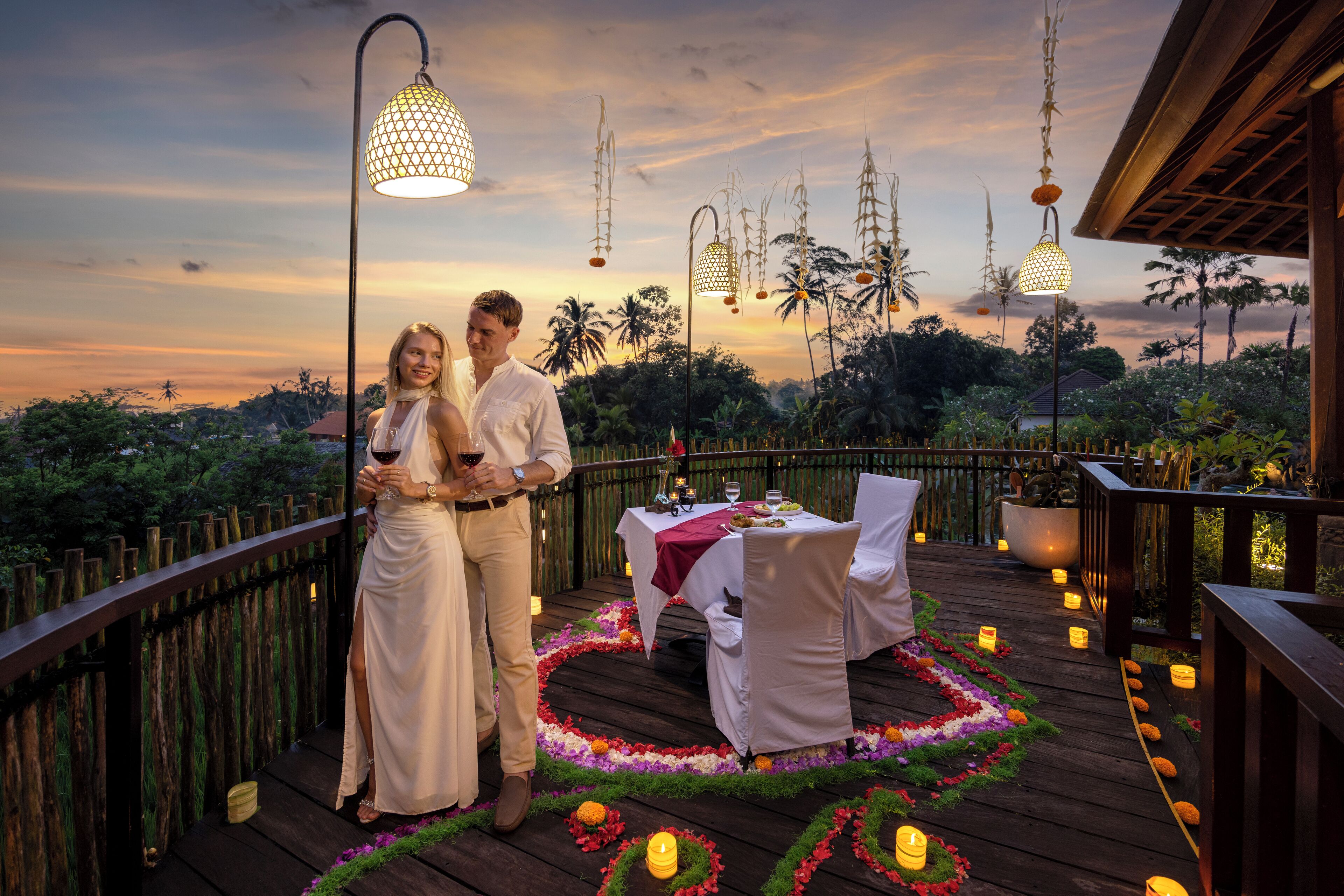 A couple standing on a wooden deck with a table set for two, surrounded by candles and flowers.