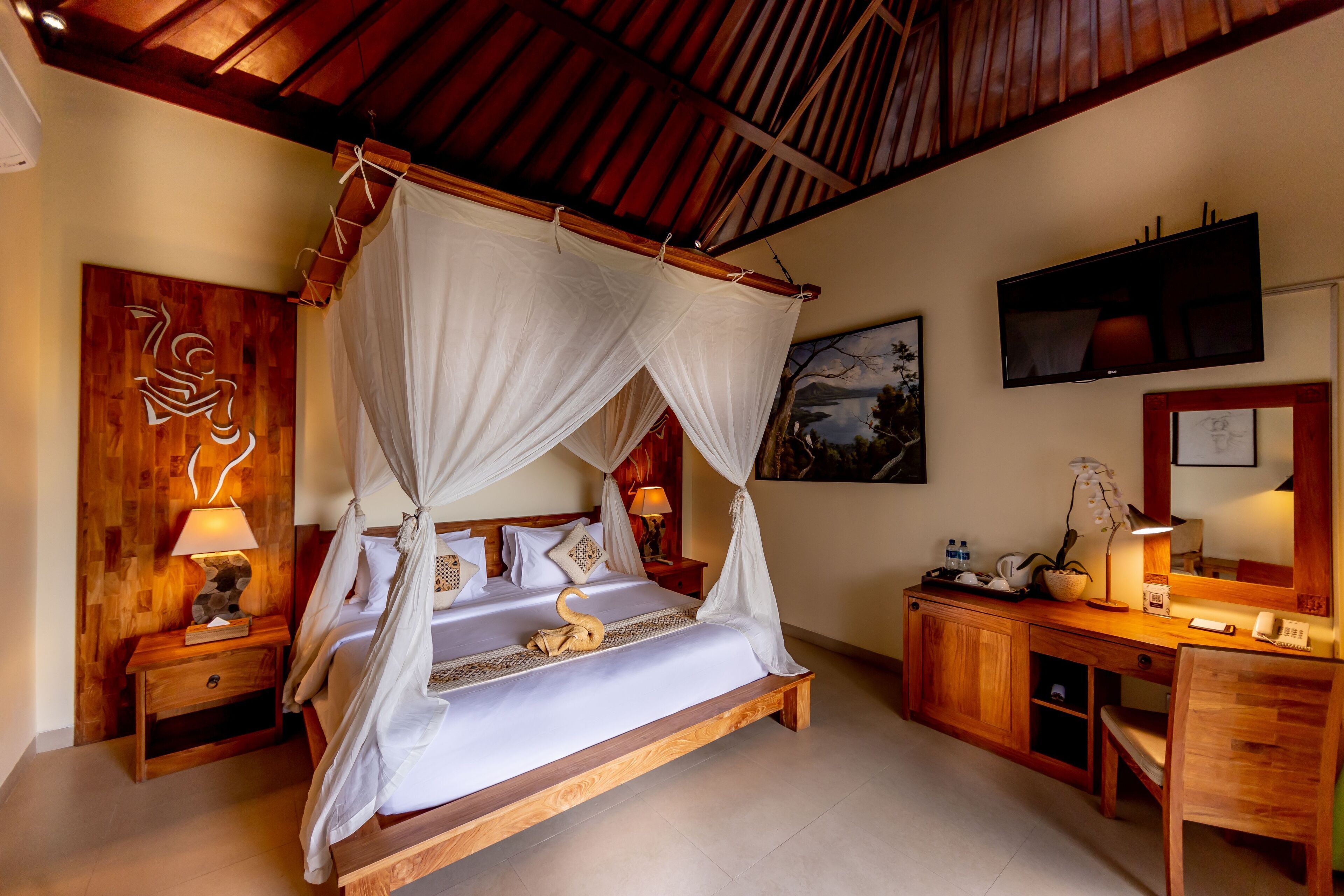 A spacious bedroom with a four-poster bed, wooden furniture, a desk with a chair, and a television on the wall.