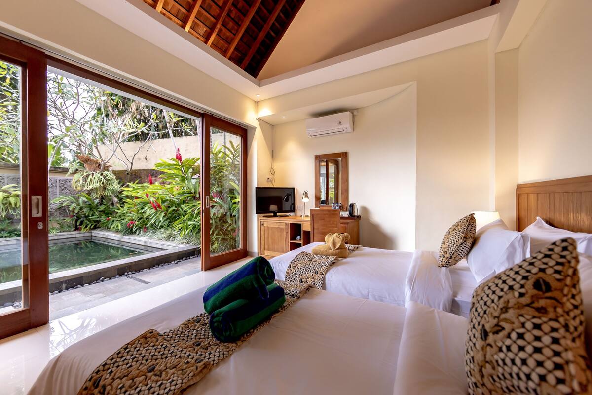 A hotel room with two beds, a TV, a desk, and a view of a garden and pool.