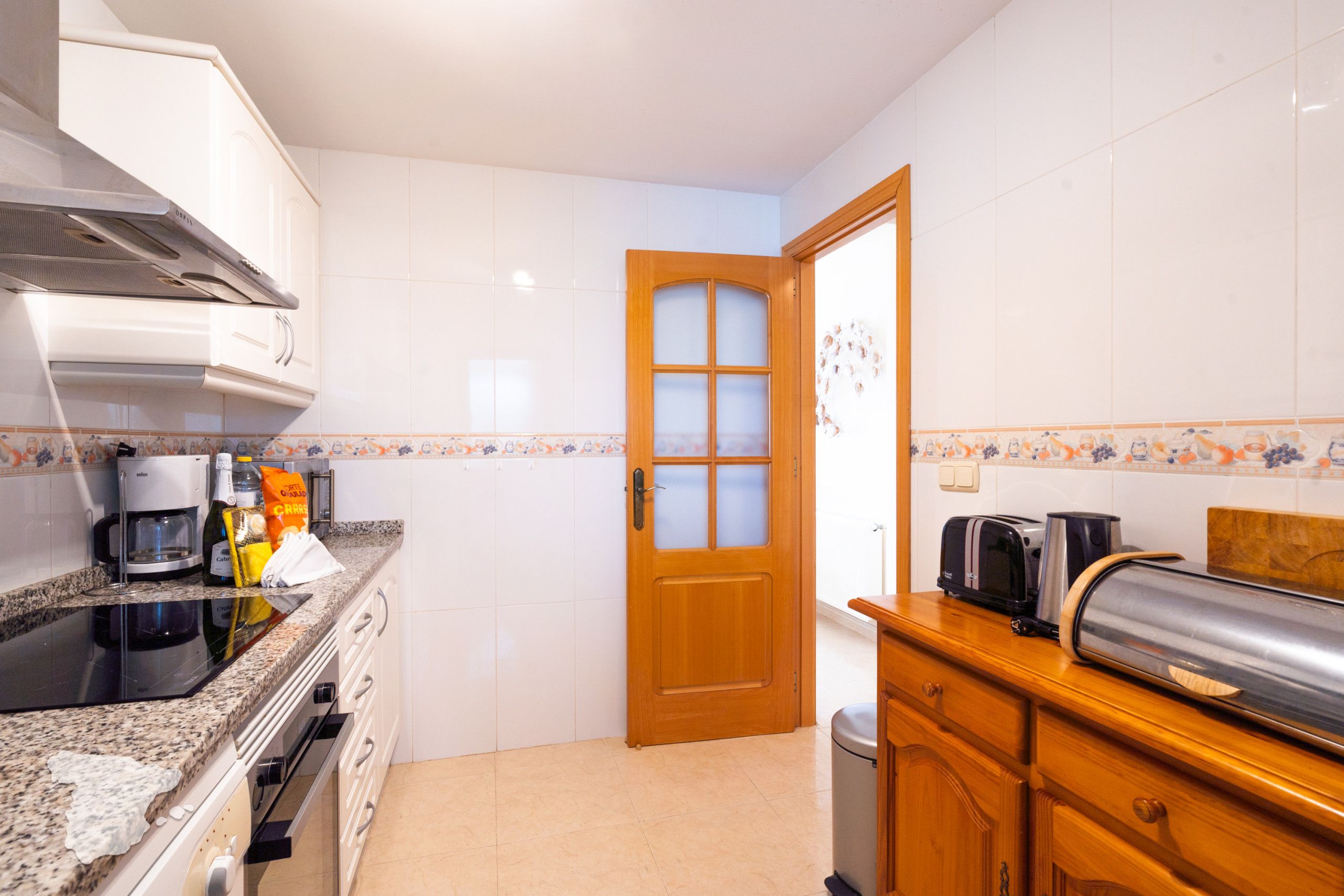 Cozinha com armários brancos, porta de madeira, cafeteira, torradeira e lixeira.