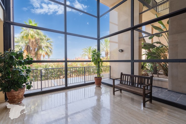 A spacious balcony with a wooden bench, potted plants, and a clear view of palm trees and flowers.