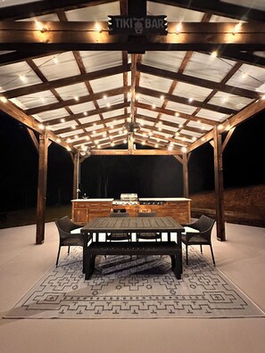 An outdoor covered patio area with a wooden table and chairs, a grill, and a sign that reads "TIKI BAR". - Ellijay Mountain Escape with Private Pool, Fire Pit, & BBQ! (Ellijay)