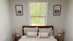 A bedroom with a large bed, two bedside tables with lamps, a window with blinds, and two framed pictures on the wall. - Ellijay Mountain Escape with Private Pool, Fire Pit, & BBQ! (Ellijay)