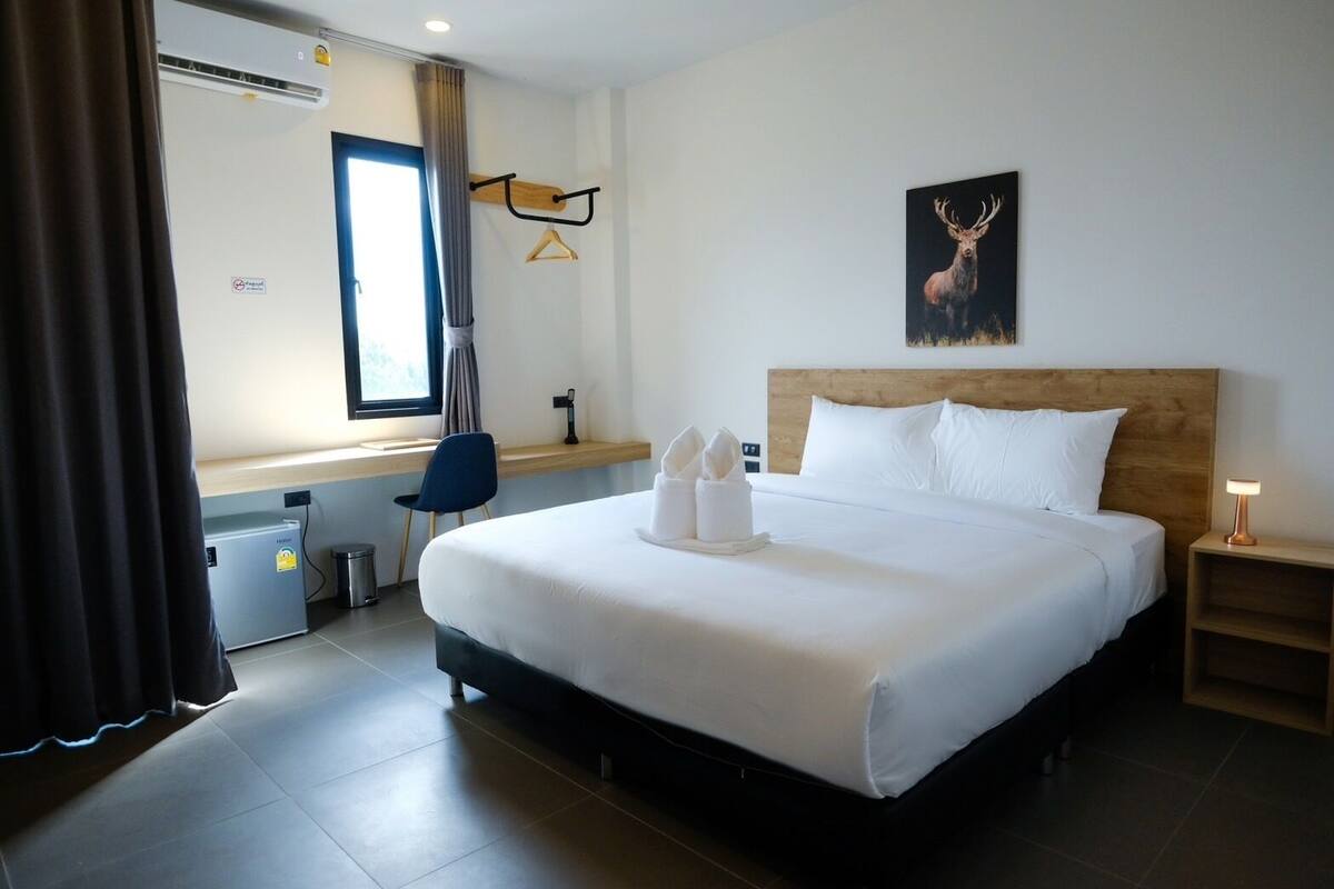 A hotel room with a large bed, a wooden headboard, a bedside table with a lamp, a window with curtains, a desk with a chair, and a wall-mounted coat rack.