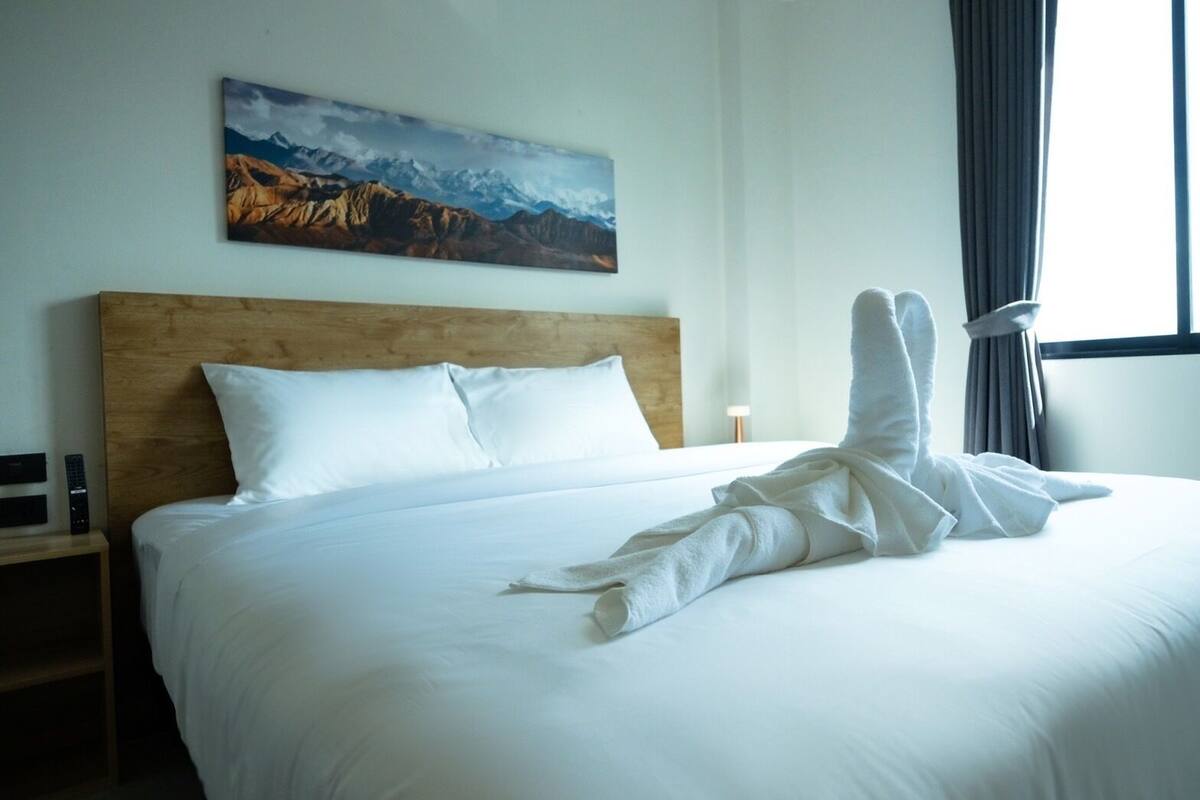 A hotel room with a large bed, a wooden headboard, a painting of mountains, and a window with curtains.