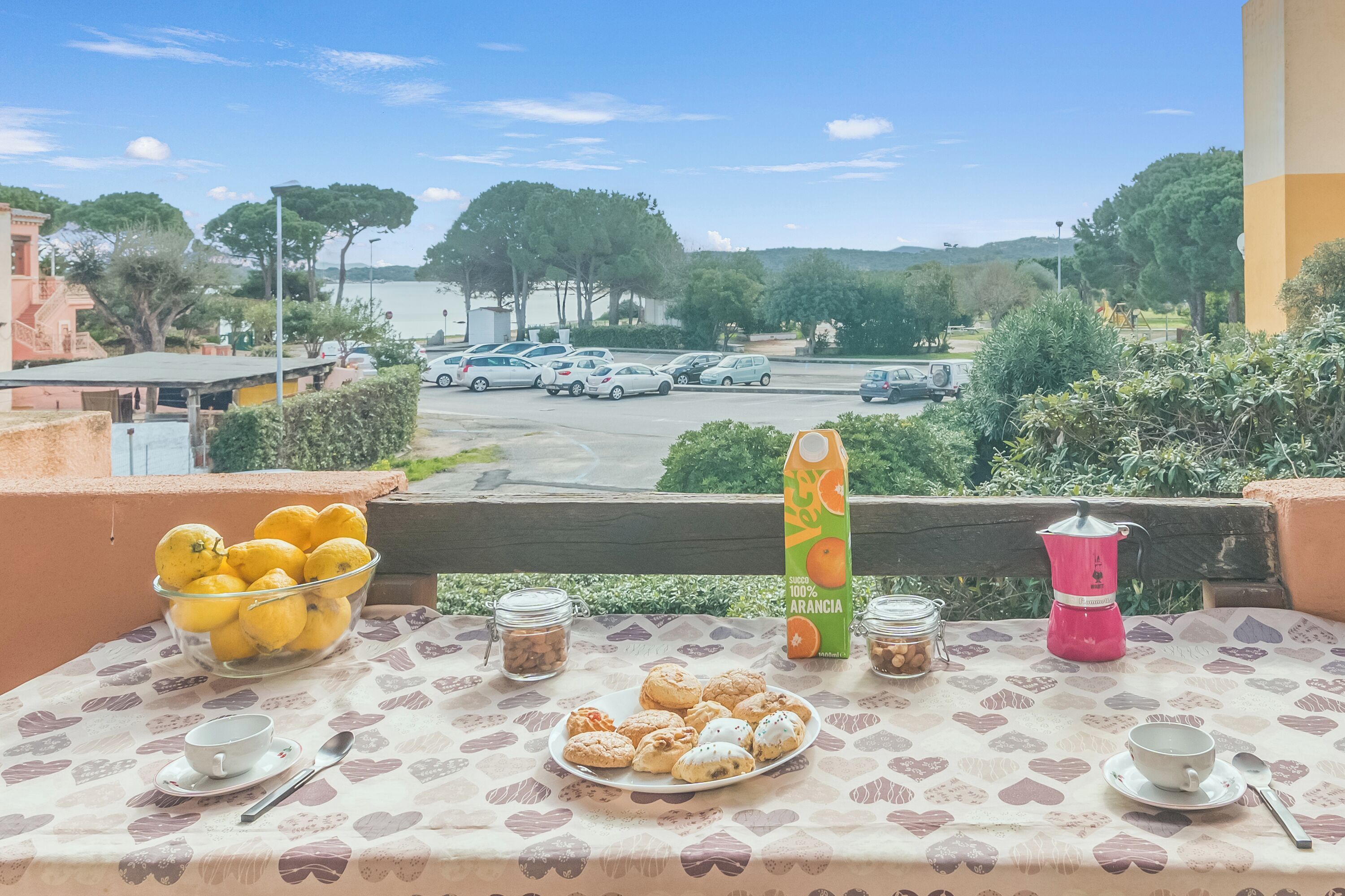A balcony with a table set for breakfast, including a bowl of lemons, a plate of pastries, and a coffee maker.