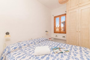 Um quarto com cama, janela, guarda-roupa de madeira, uma mesinha e uma luminária.