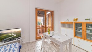 Ein kleiner, heller Raum mit Fernseher, Esstisch, Stühlen, einem Schrank und Blick auf einen Garten durch eine Tür.