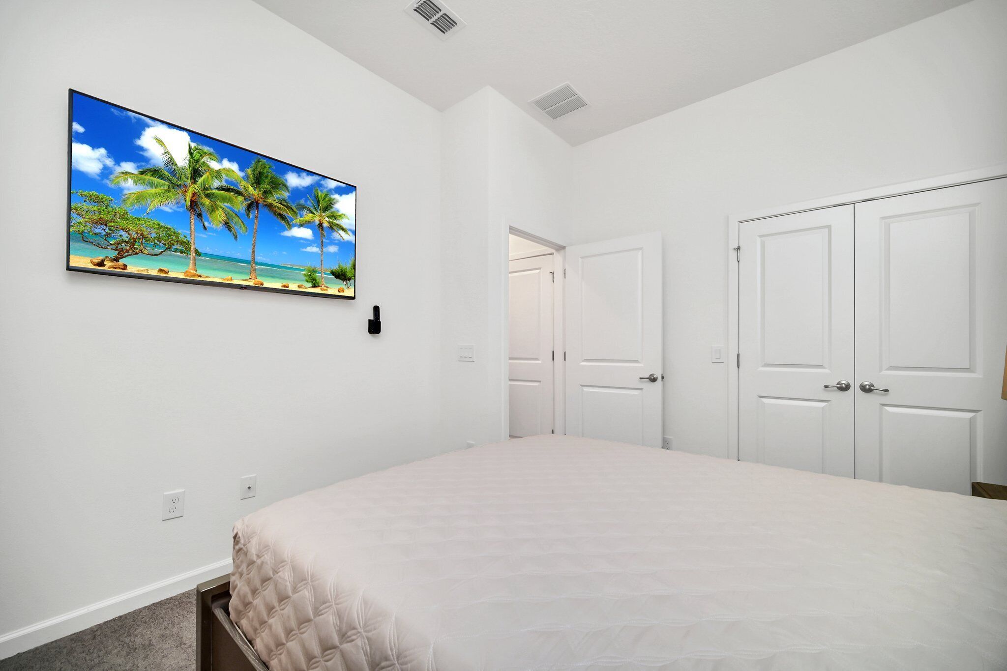 Un dormitorio con una cama, paredes blancas, un cuadro de una playa tropical y puertas blancas.