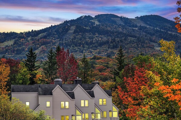 A two-story house with multiple windows, surrounded by autumn foliage and a mountainous backdrop.