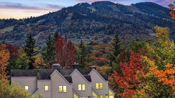 A two-story house with multiple windows and a sloped roof, surrounded by autumn foliage and a mountain backdrop. - Beautiful 2BR/2BA Fall Line Condo w/ Mountain Views, Pool, Hot Tub, Sauna, Shuttle. G6B (Killington)