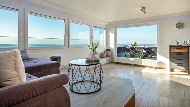 A modern living room with a sofa, a coffee table, and a view of the sea through large windows. - 1-room penthouse with rooftop terrace, stunning lake & mountain views, and green surroundings (Tettnang)