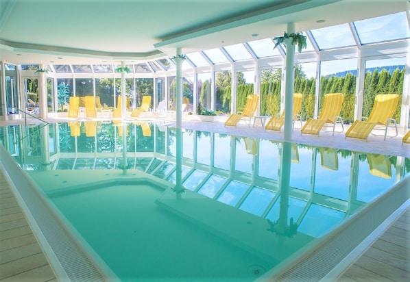 Une piscine intérieure avec des parois en verre transparent, des chaises longues jaunes et une vue sur les arbres et les montagnes.