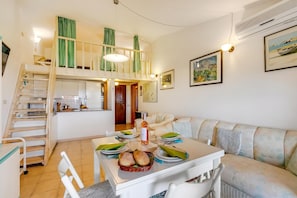 Zona pranzo con tavolo apparecchiato per due, divano, una scala e cucina sullo sfondo.