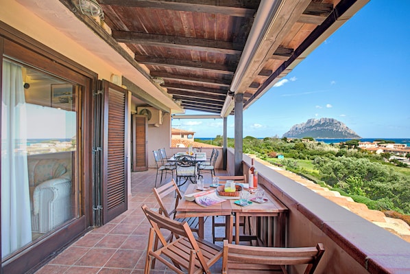 A covered outdoor dining area with wooden furniture and a view of a mountain and sea.