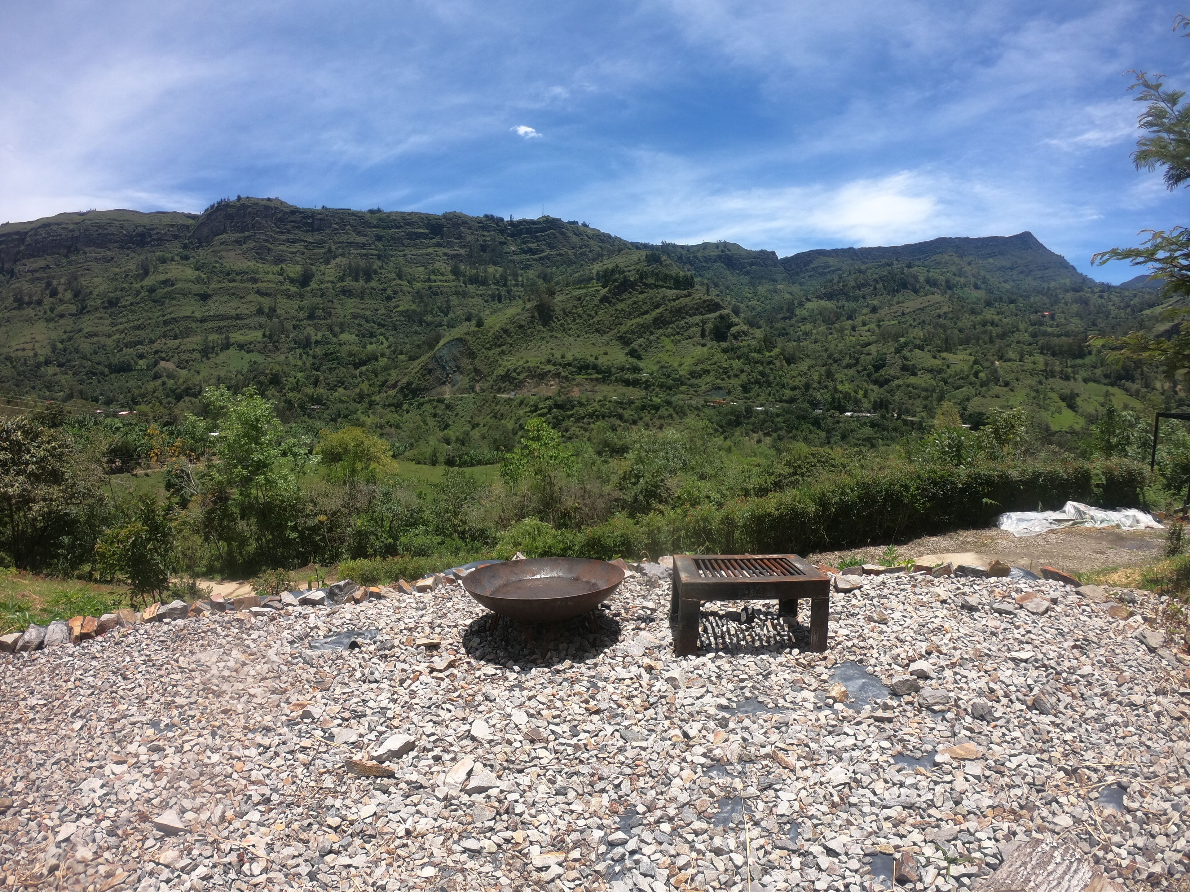A rocky outdoor area with a metal fire pit and a wooden bench, surrounded by a mountainous landscape.