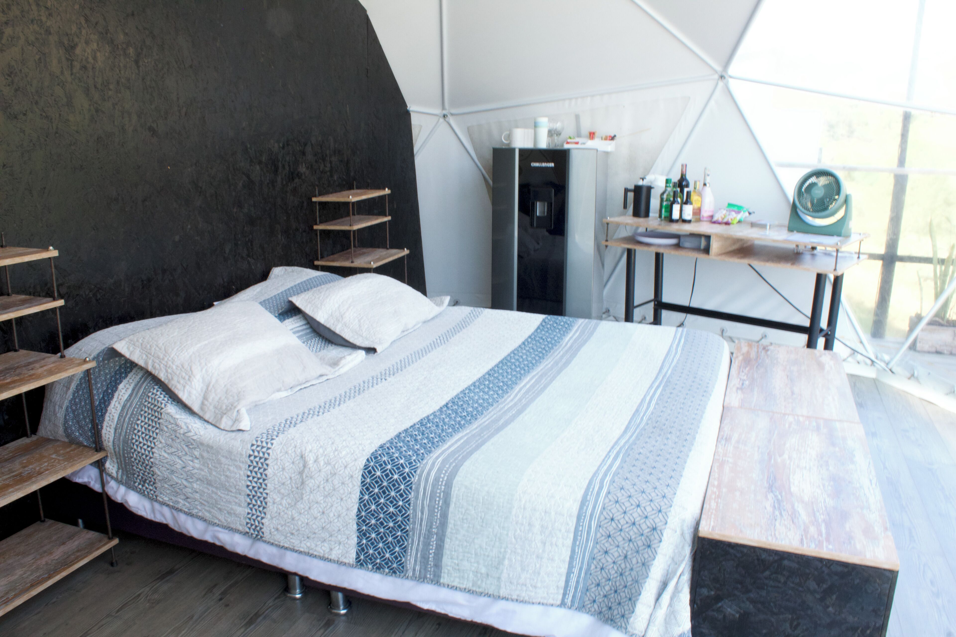 A bed with a patterned blanket and pillows, a wooden bench, a small table with bottles, and a water dispenser in a tent-like room.