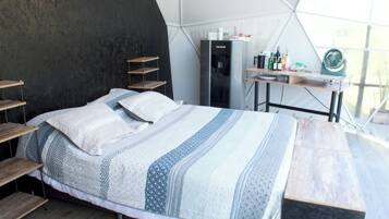 A bed with a patterned blanket and pillows, a wooden bench, a small table with bottles, and a water dispenser in a tent-like room.