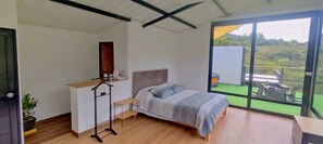 Un dormitorio con cama, escritorio, silla, planta en maceta y ventana con vistas a un paisaje verde.