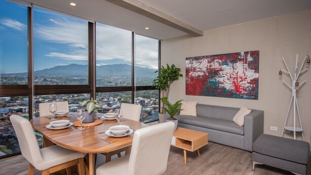 A modern living room with a dining area, large windows with a city view, a gray sofa, and a red abstract painting. - Digital Nomads Dream + Sauna (Curridabat)