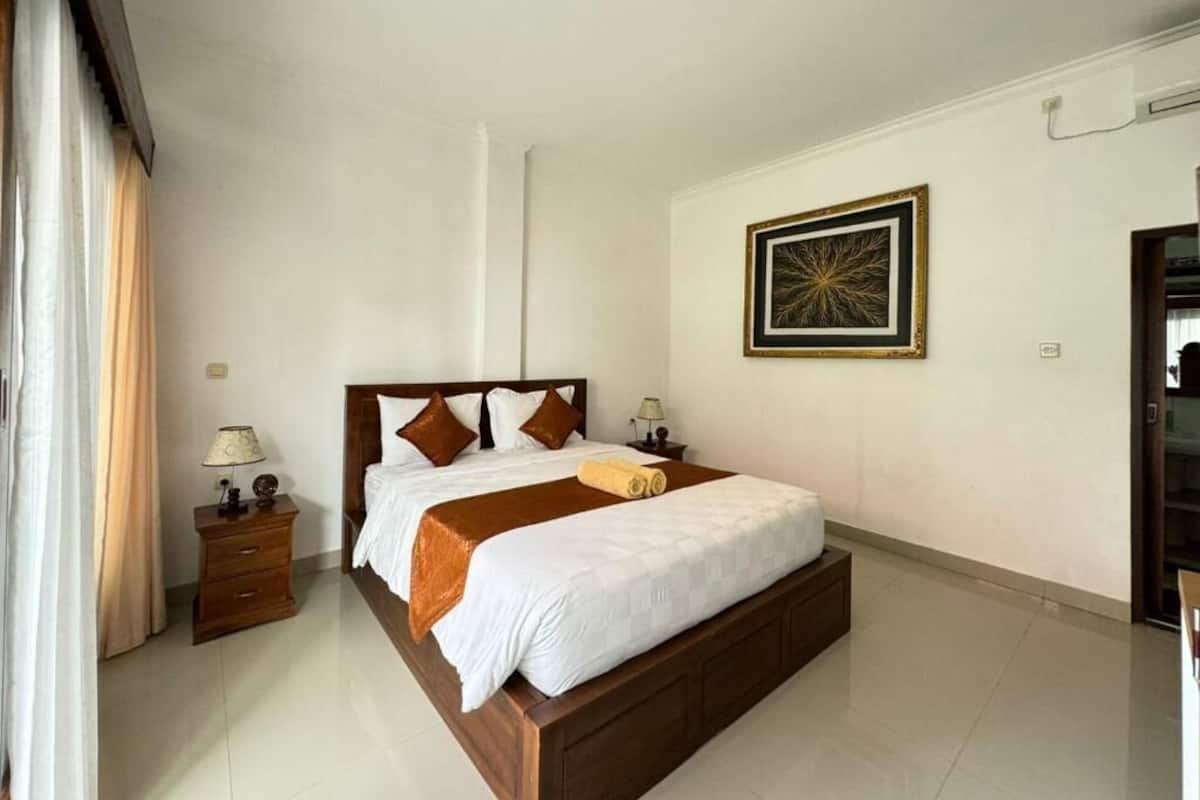 A bedroom with a large bed, bedside tables, a wall-mounted artwork, and a door to the right.