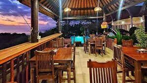 An outdoor dining area with wooden tables and chairs, a pool in the background, and a thatched roof structure. - Kaylas Hill Resort (Penida Island)