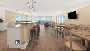 A spacious kitchen with a central island, a dining area with wooden tables and chairs, and a large TV mounted on the wall.