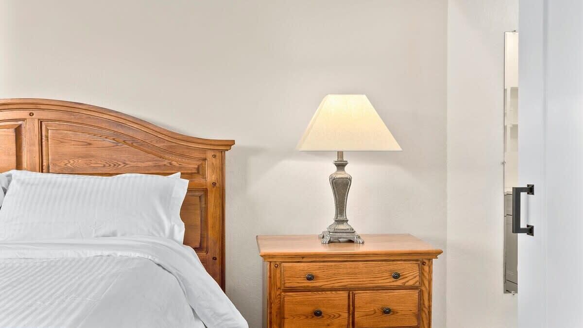 Una cama de madera con edredón blanco, una mesita de noche con lámpara y una puerta a la derecha.