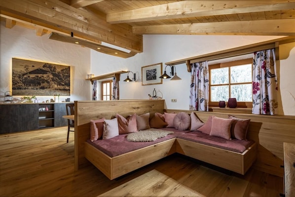 A cozy wooden interior with a L-shaped sofa, wooden beams, a kitchen area, and large windows with floral curtains.