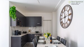Una cucina moderna con mobili scuri, un forno a microonde e una macchina per il caffè. C'è un'area pranzo con un tavolo apparecchiato per quattro persone, una sveglia e un vaso con fiori.
