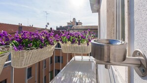 Un balcone con cesti in vimini pieni di fiori viola, una pentola in acciaio inossidabile e vista su edifici sullo sfondo.