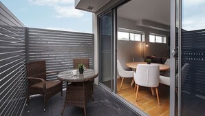 Un balcón moderno con mobiliario de mimbre, puerta de vidrio que da a un comedor interior con sillas blancas y una mesa redonda.
