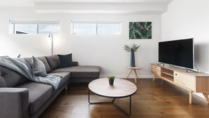 Una sala de estar moderna con un sofá gris, una mesita redonda, un televisor sobre un mueble de madera y una planta en una mesa pequeña.
