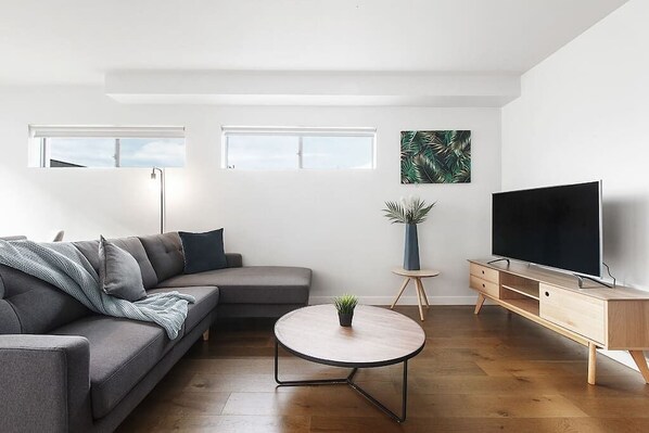 A modern living room with a grey sofa, a round coffee table, a TV on a wooden stand, and a plant on a small table.