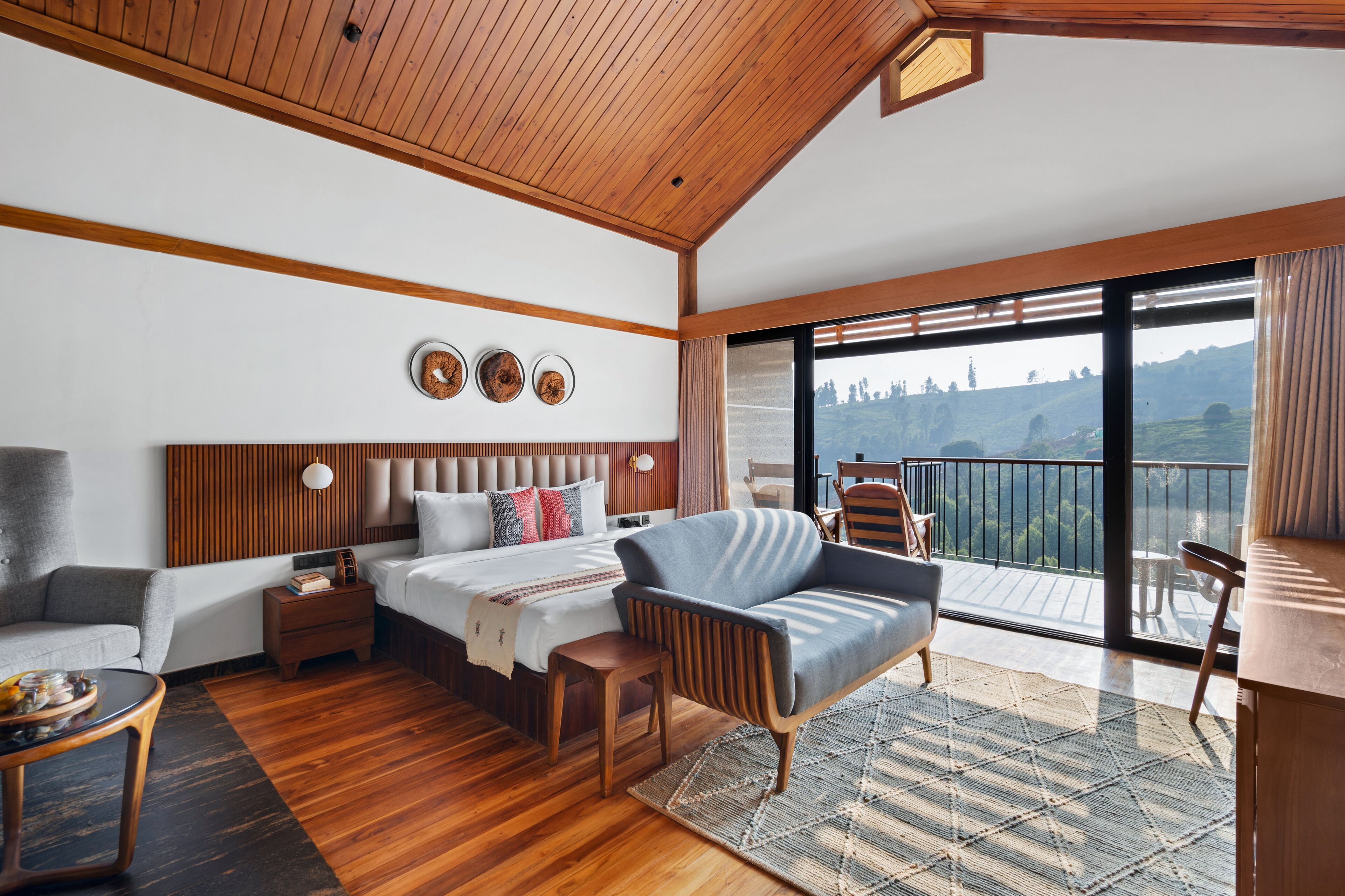 A modern hotel room with a wooden ceiling, a large bed with a headboard, a sofa, a small table, and a balcony with a view of the hills.