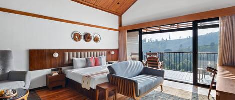 A modern hotel room with a wooden ceiling, a large bed with a headboard, a sofa, a small table, and a balcony with a view of the hills.