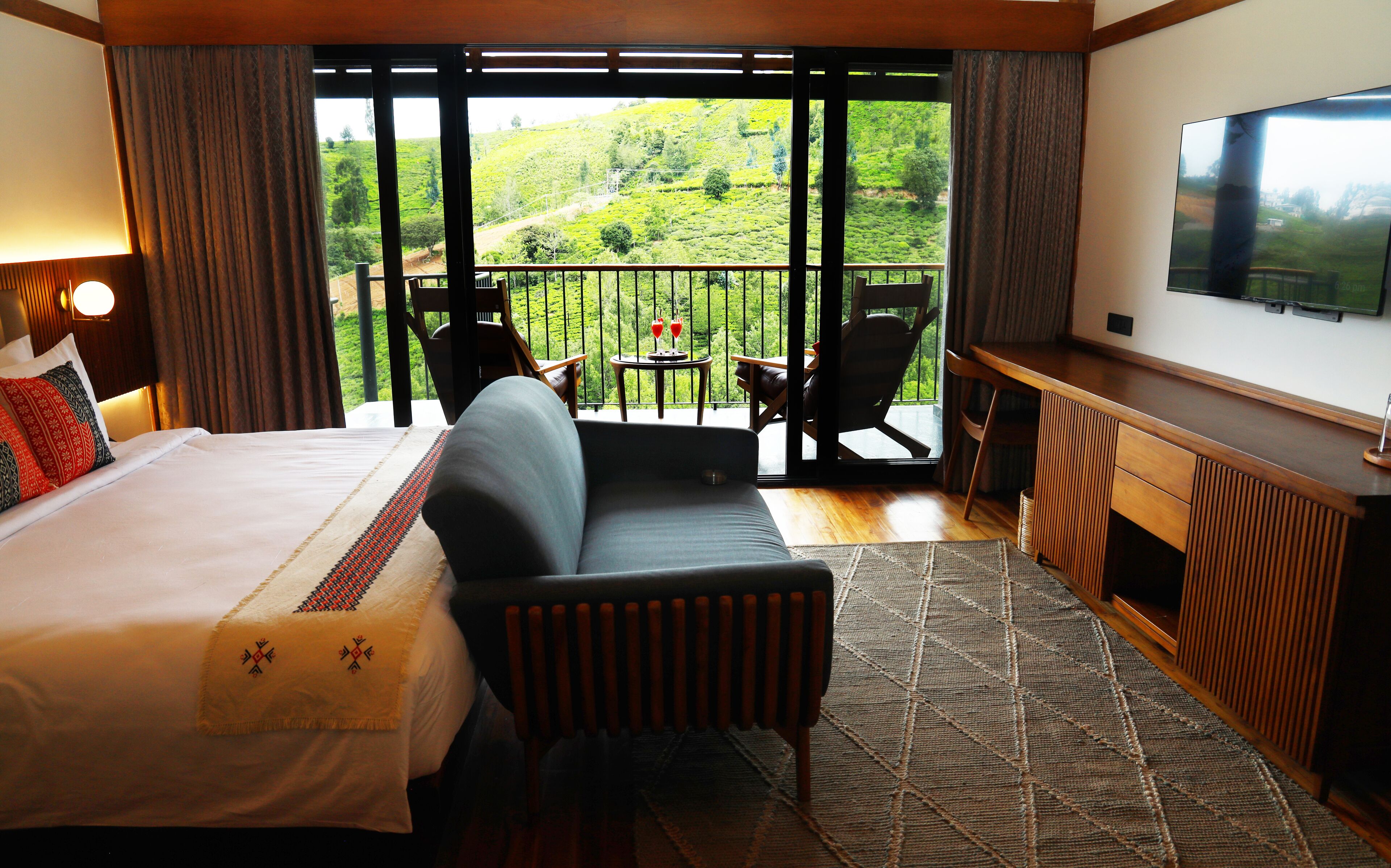 A hotel room with a bed, a sofa, a desk, and a TV. The room has a balcony with a view of green hills.
