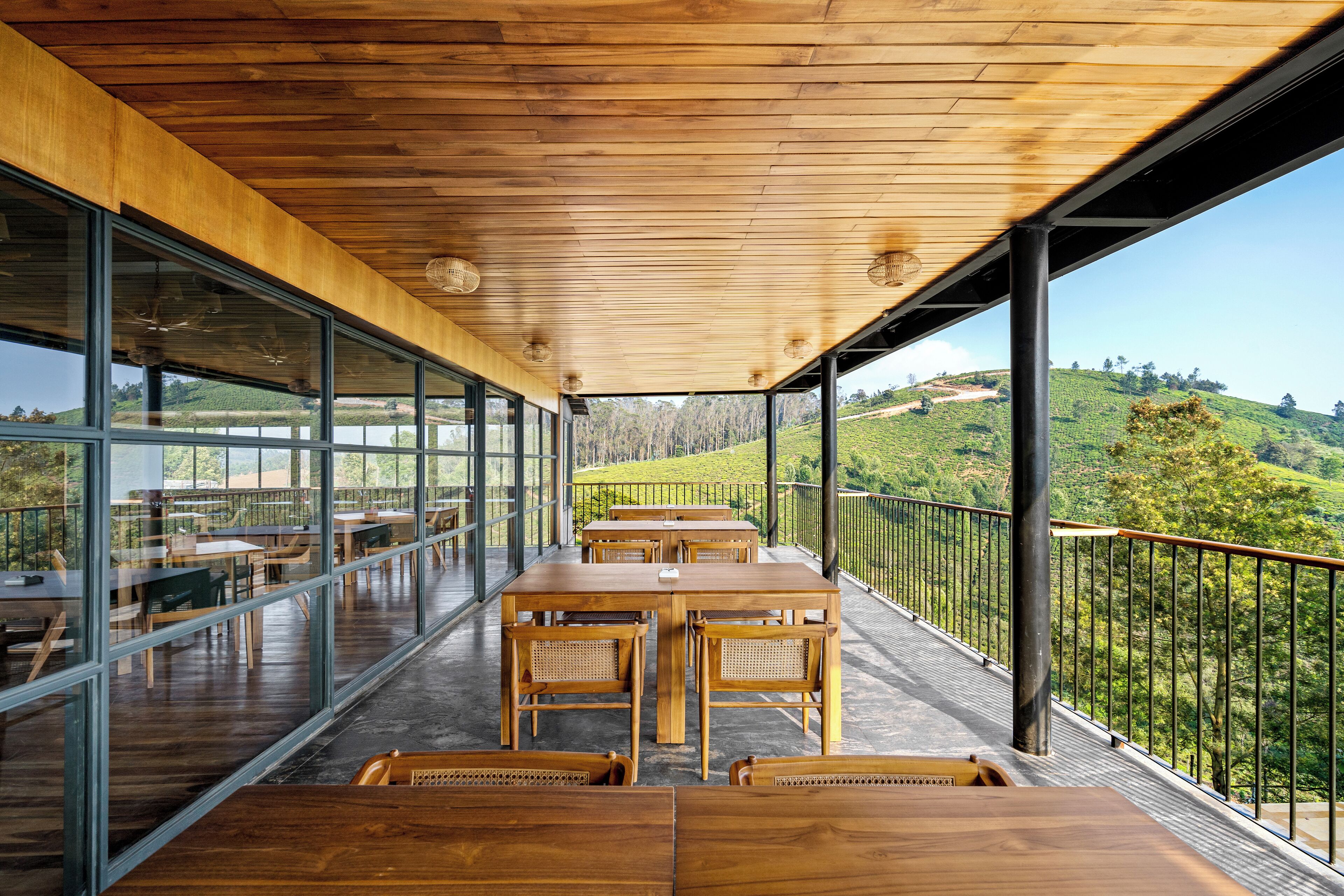 Um terraço amplo ao ar livre, com mesas e cadeiras de madeira, grandes janelas de vidro e uma vista panorâmica das colinas.