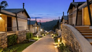 A pathway leading to a row of modern, light-colored buildings with stone accents, surrounded by greenery and set against a hilly backdrop. - WelcomHeritage Ayatana Ooty (Coonoor)