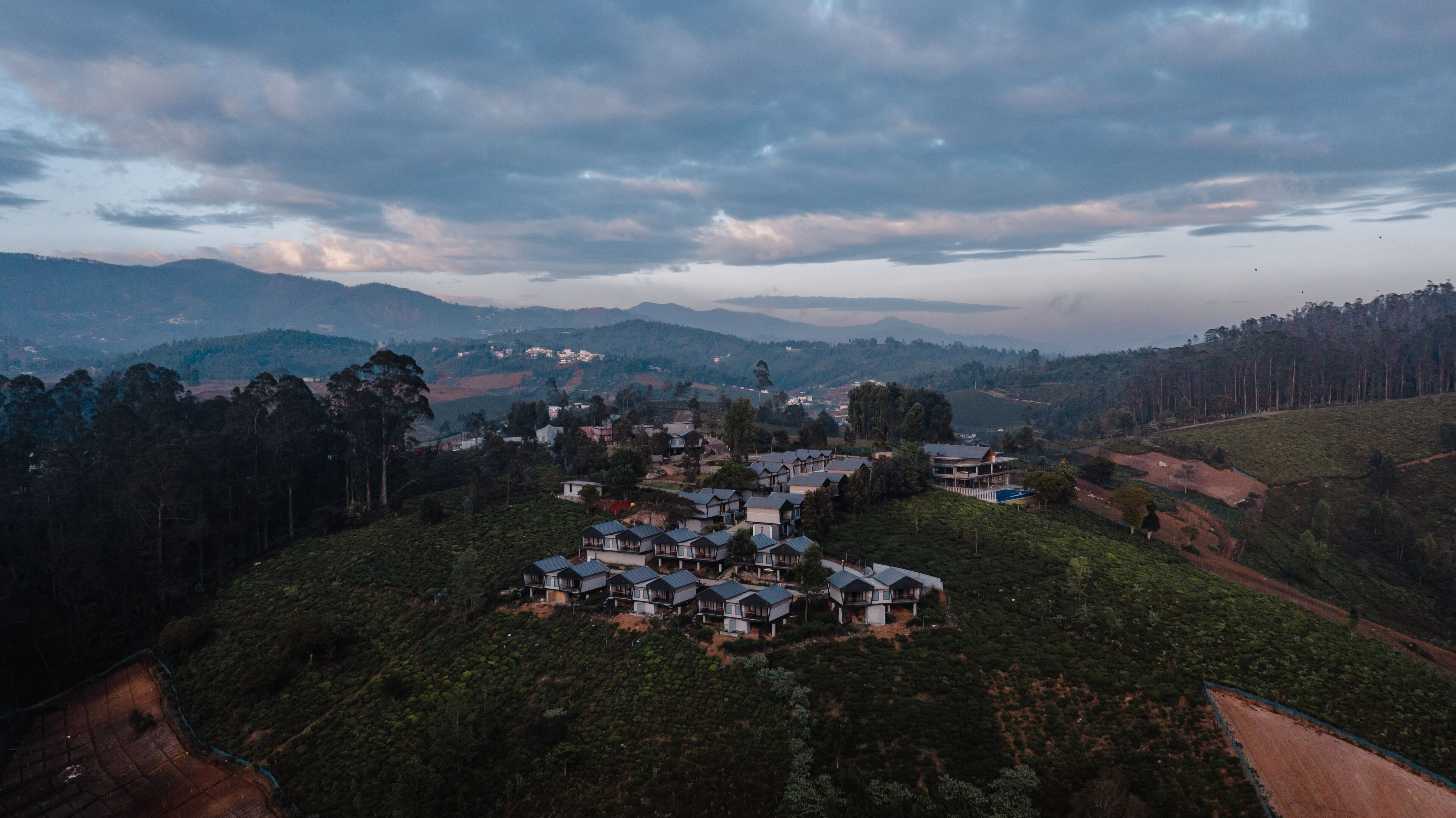 A residential area with multiple houses on a hillside, surrounded by dense forest and rolling hills in the background.