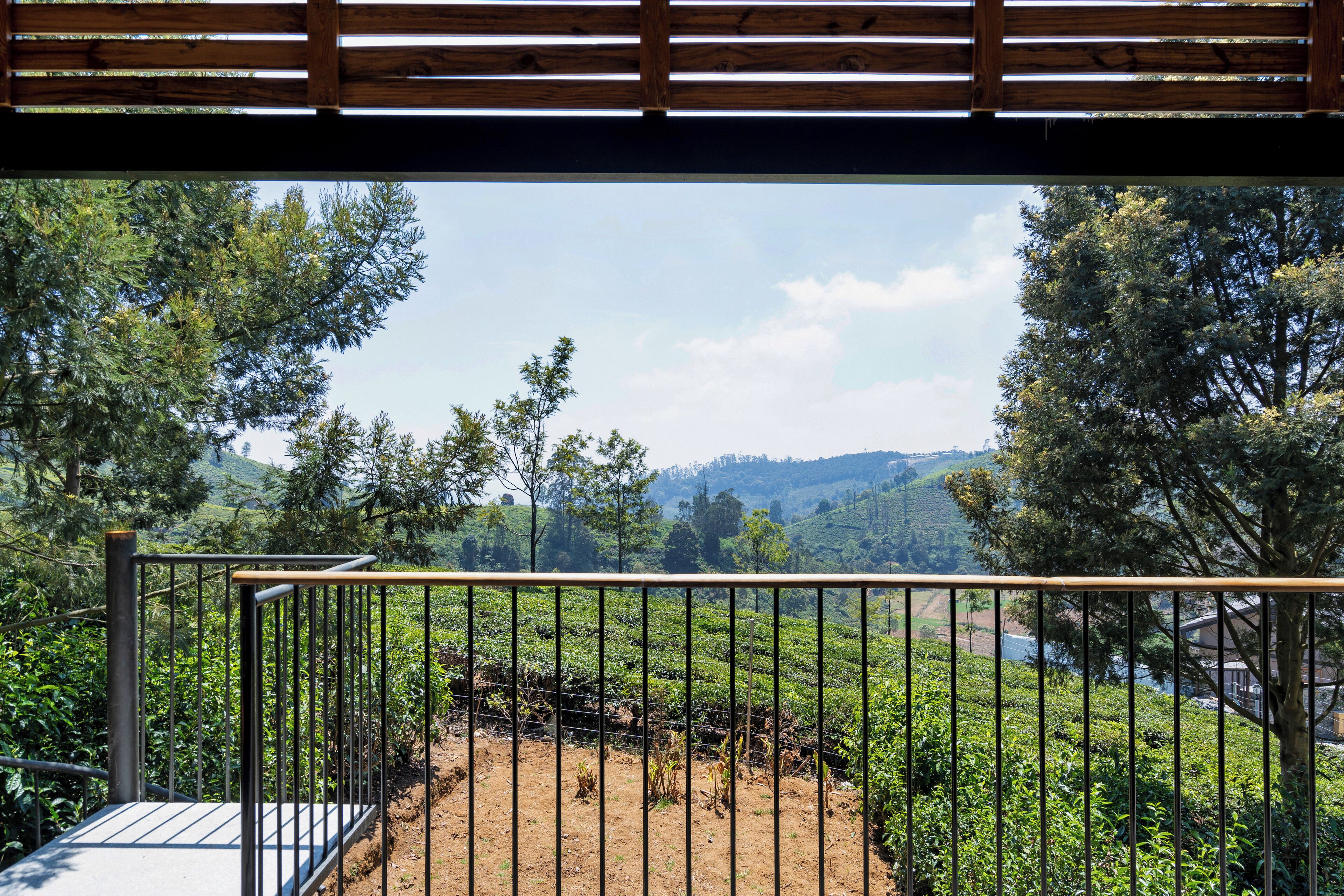 A view from a balcony with a metal railing, overlooking a lush landscape with trees and a distant hill.