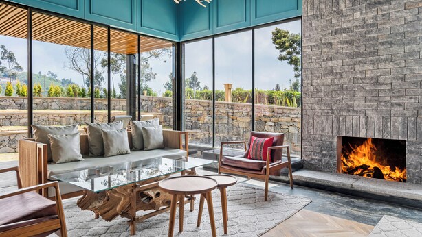 A modern living room with a glass coffee table, wooden chairs, a sofa with cushions, and a fireplace. - WelcomHeritage Ayatana Ooty (Coonoor)