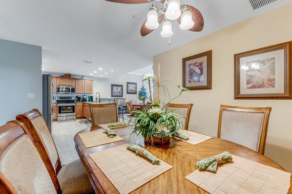 Un coin repas avec une table en bois dressée pour quatre personnes, entourée de chaises. On peut voir un ventilateur de plafond et des lampes fixées au mur.