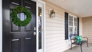 A front porch with a black door, a green wreath, a bench with a cushion, and a wall-mounted lantern.