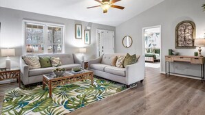A living room with a white sofa, a coffee table, and a ceiling fan. There are windows, a mirror, and a door visible.
