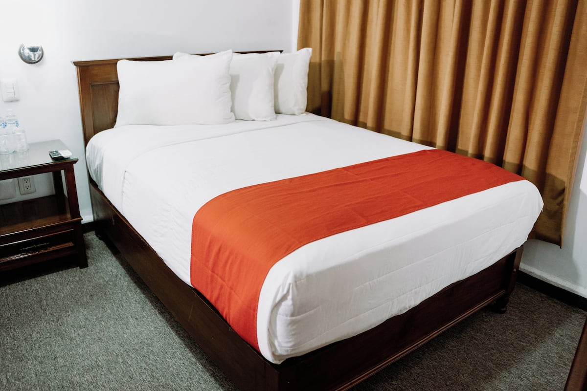 A neatly made bed with a white and red bedspread, a wooden headboard, and a nightstand with a glass of water and a remote control.
