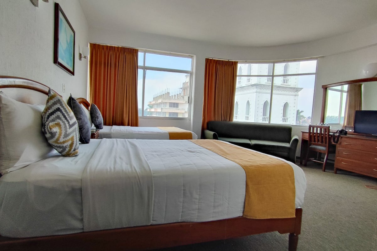 A hotel room with two beds, a desk, a chair, and large windows with curtains.