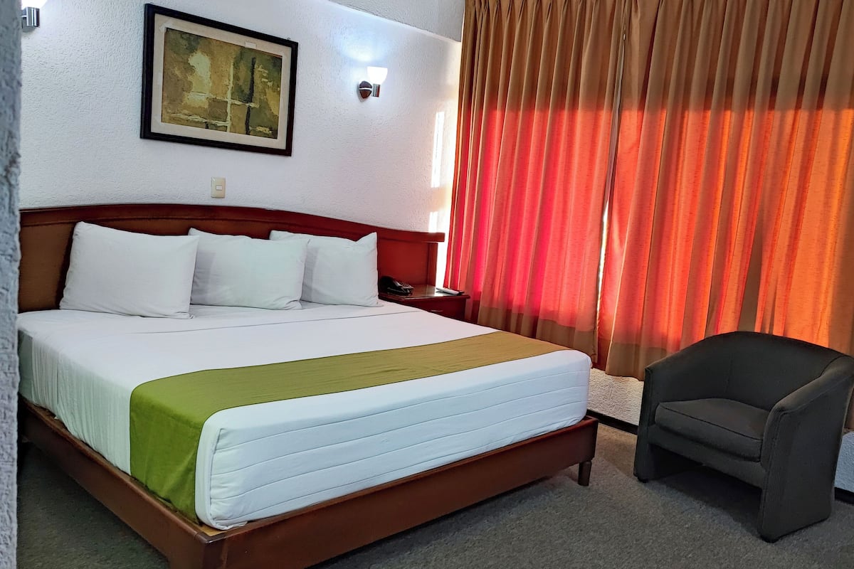 A hotel room with a large bed, a chair, a painting on the wall, and a window with curtains.